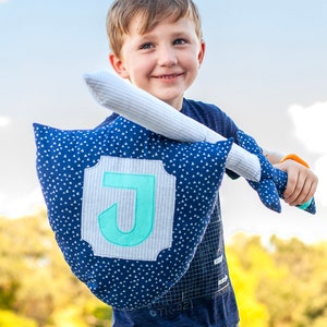 May include: A young boy holds a blue and white pillow with a large, light blue letter J in the center. The pillow is shaped like a shield and has a white sword attached to it.