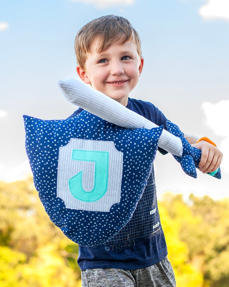 May include: A young boy holds a blue and white pillow with a large, light blue letter J in the center. The pillow is shaped like a shield and has a white sword attached to it.