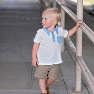 May include: A young boy wearing a white polo shirt with a blue collar and a blue pocket. He is wearing khaki shorts and brown sandals. He is standing in a walkway with a metal railing.