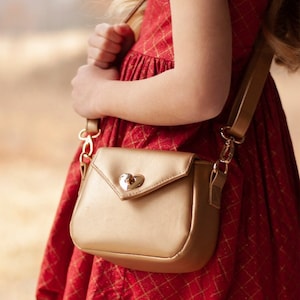 May include: A gold-colored faux leather crossbody bag with a heart-shaped clasp. The bag has a flap closure and a long adjustable strap.