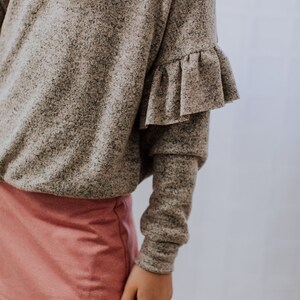 May include: A gray speckled long-sleeve top with ruffled sleeves and a pink skirt. The top has a loose fit, and the skirt is a solid color. The outfit is casual and comfortable.