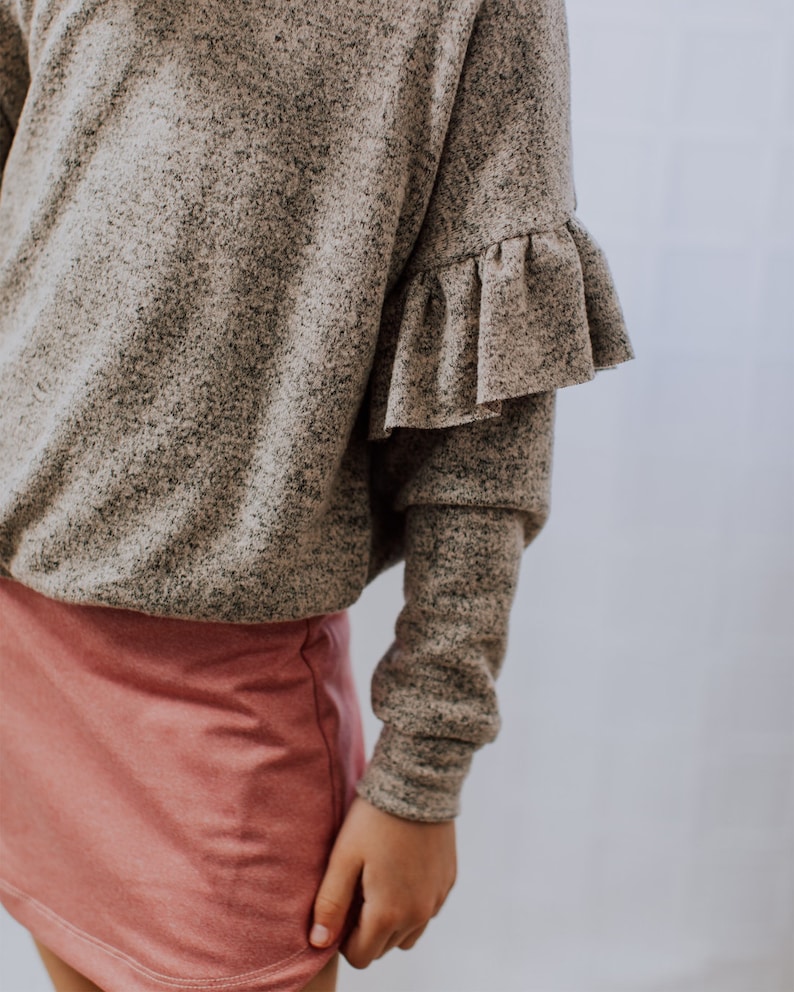 May include: A gray speckled long-sleeve top with ruffled sleeves and a pink skirt. The top has a loose fit, and the skirt is a solid color. The outfit is casual and comfortable.