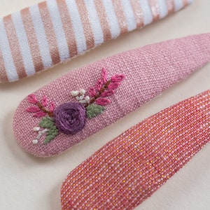 Pu&ograve; includere: Tre fermagli per capelli in tessuto rosa. Un fermaglio ha un motivo a righe bianche e dorate, un altro ha un motivo a quadri rosa e dorati e l'ultimo ha un ricamo floreale rosa.