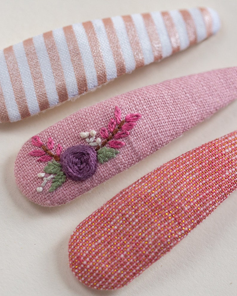Pu&ograve; includere: Tre fermagli per capelli in tessuto rosa. Un fermaglio ha un motivo a righe bianche e dorate, un altro ha un motivo a quadri rosa e dorati e l'ultimo ha un ricamo floreale rosa.