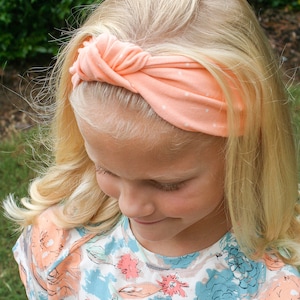 Könnte beinhalten: Ein junges Mädchen mit blonden Haaren trägt ein pfirsichfarbenes Stirnband mit weißen Punkten. Das Stirnband ist oben auf ihrem Kopf zu einem Knoten gebunden. Sie trägt ein Hemd mit Blumenmuster.