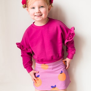 May include: A young child wearing a bright pink sweatshirt with ruffled sleeves and a matching pink skirt. The skirt features a pattern of orange fruit with blue leaves. The child is smiling and has pink hair accessories.