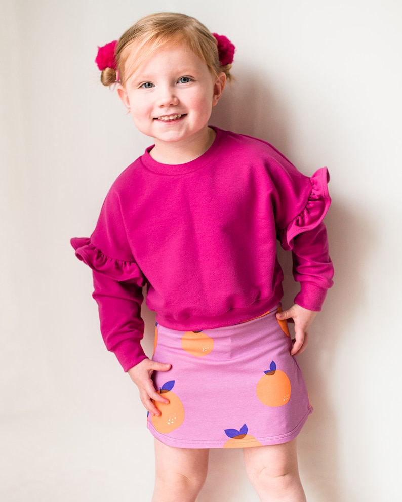 May include: A young child wearing a bright pink sweatshirt with ruffled sleeves and a matching pink skirt. The skirt features a pattern of orange fruit with blue leaves. The child is smiling and has pink hair accessories.