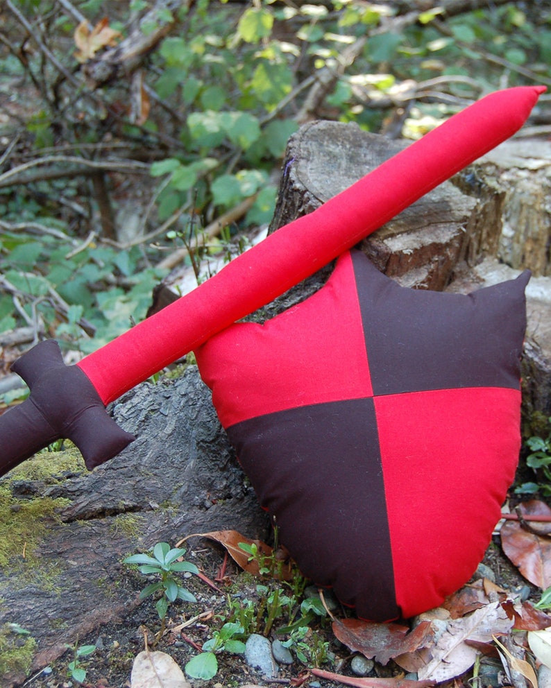 May include: A red and black fabric sword and shield set. The sword is made of fabric and has a red handle. The shield is also made of fabric and has a red and black checkered pattern.
