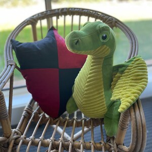 May include: A green plush dragon with yellow wings sits on a wicker chair with a black and red checkered pillow.