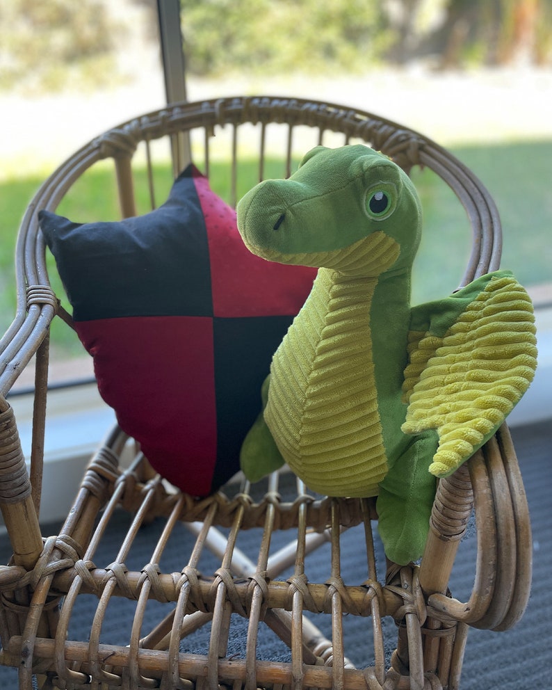 May include: A green plush dragon with yellow wings sits on a wicker chair with a black and red checkered pillow.