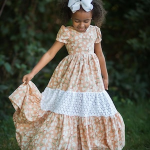 May include: A peach-colored floral dress with a white lace panel. The dress has short sleeves and a full skirt. A white bow is in the hair. The dress is suitable for a special occasion or everyday wear.