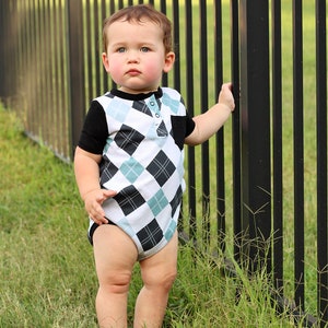 Może przedstawiać: Małe dziecko w biało-czarnej koszuli w argyle z niebiesko-białym wzorem w romby. Dziecko stoi obok czarnego metalowego ogrodzenia.