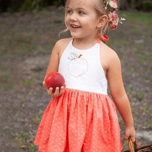 K&ouml;nnte beinhalten: Ein junges M&auml;dchen tr&auml;gt ein wei&szlig;es Halfterkleid mit einem Pfirsich-Stickerei-Design und einen pfirsichfarbenen Rock mit Blumenmuster. Sie h&auml;lt einen Pfirsich in der Hand und einen Weidenkorb.