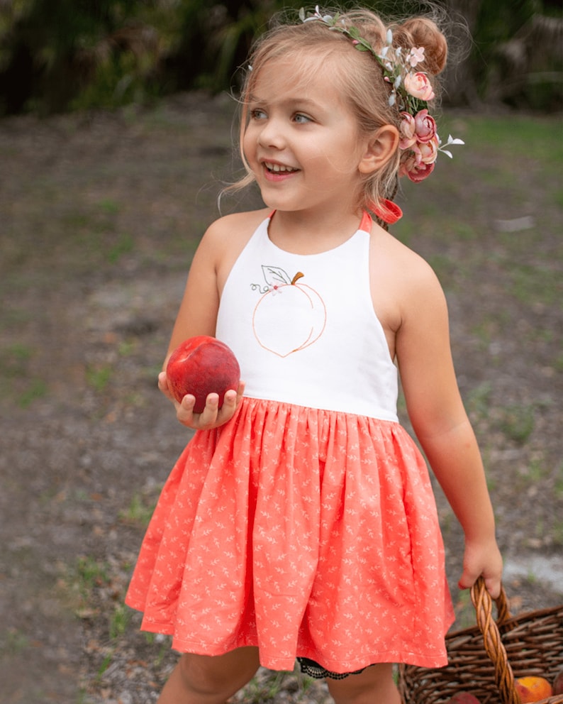 K&ouml;nnte beinhalten: Ein junges M&auml;dchen tr&auml;gt ein wei&szlig;es Halfterkleid mit einem Pfirsich-Stickerei-Design und einen pfirsichfarbenen Rock mit Blumenmuster. Sie h&auml;lt einen Pfirsich in der Hand und einen Weidenkorb.