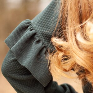 May include: Close-up of a dark teal green sleeve with a textured pattern and a ruffled cuff. The sleeve is part of a garment, possibly a dress or blouse. Golden blonde hair is visible in the background, adding contrast to the dark fabric.