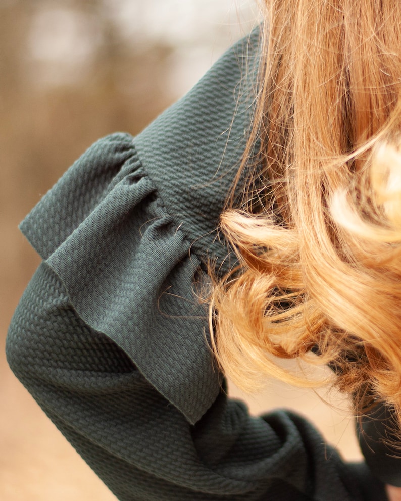 May include: Close-up of a dark teal green sleeve with a textured pattern and a ruffled cuff. The sleeve is part of a garment, possibly a dress or blouse. Golden blonde hair is visible in the background, adding contrast to the dark fabric.