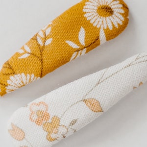 Pu&ograve; includere: Due fermagli per capelli in tessuto con motivi floreali. Un fermaglio &egrave; giallo senape con fiori e foglie bianche. L'altro fermaglio &egrave; bianco con fiori e foglie di colore arancio chiaro e marrone.