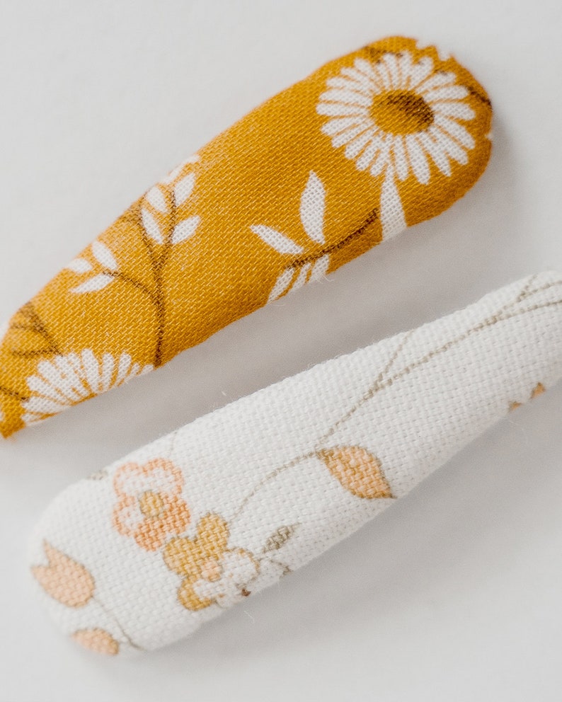 Pu&ograve; includere: Due fermagli per capelli in tessuto con motivi floreali. Un fermaglio &egrave; giallo senape con fiori e foglie bianche. L'altro fermaglio &egrave; bianco con fiori e foglie di colore arancio chiaro e marrone.