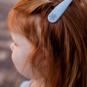 Pu&ograve; includere: Un fermaglio per capelli blu chiaro con un design a fiocco di neve argentato. Il fermaglio &egrave; nei capelli rossi di una ragazza.