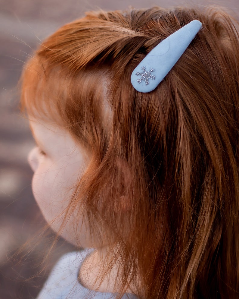 Pu&ograve; includere: Un fermaglio per capelli blu chiaro con un design a fiocco di neve argentato. Il fermaglio &egrave; nei capelli rossi di una ragazza.
