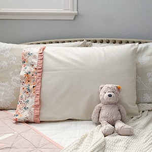 Puede incluir: Una almohada blanca con un volante de estampado floral en el borde. La almohada está en una cama con una colcha rosa y blanca y una manta de punto blanca. Un oso de peluche gris está sentado en la manta.