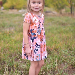 May include: A young girl wearing a pink and orange floral dress with short sleeves. The dress has a white background with a variety of colored floral patterns. The girl is standing in a field of green grass.