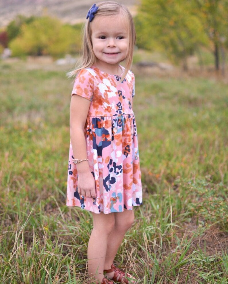 May include: A young girl wearing a pink and orange floral dress with short sleeves. The dress has a white background with a variety of colored floral patterns. The girl is standing in a field of green grass.