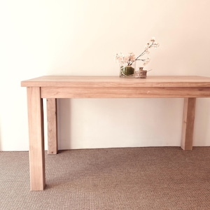 May include: A rectangular light-toned wooden table with four straight legs. On the table surface sits a clear glass vase containing delicate pale pink cherry blossom branches and a small, round, lidded wooden container next to the vase.