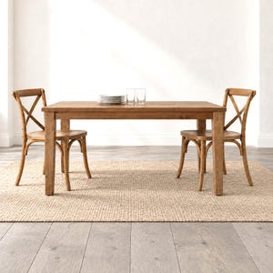 May include: A rectangular wooden dining table with four straight legs and two matching wooden chairs featuring an X-shaped back design. The table surface holds a stack of white plates and two clear drinking glasses, all set on a woven rug.
