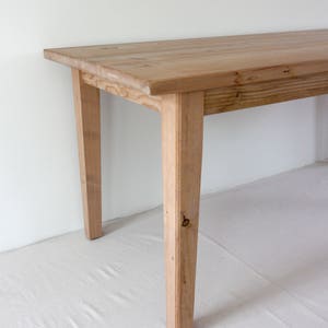 May include: A rectangular wooden table featuring a light brown natural wood grain finish. The table has four straight, tapered legs and a smooth, flat top surface. The construction shows visible wood knots and a clear, light-toned timber texture.