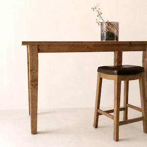 May include: A rectangular wooden table with a natural brown finish and four straight legs. Beside the table stands a wooden stool featuring a dark brown padded seat. A glass vase with a dark spiral pattern and small white flowers sits on top.