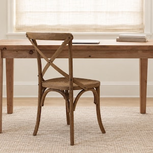 May include: A light brown wooden desk with four straight legs and a matching wooden chair featuring a cross-back design and a woven seat. The desk surface is rectangular and holds a thin laptop and a small stack of books.