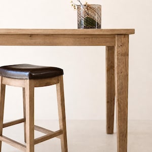 May include: A tall wooden table with a natural light brown finish and a matching wooden stool featuring a dark brown leather seat. A cylindrical glass vase with a dark swirling pattern and small white flowers sits on the table surface.