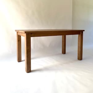 May include: A rectangular wooden table with a medium brown finish, featuring a flat tabletop surface and four sturdy, straight legs. The table is constructed with visible wood grain patterns throughout the frame and legs, showcasing a simple design.