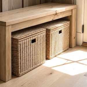 May include: A light-toned wooden bench with a flat rectangular seat and four sturdy legs. Underneath the bench are two rectangular woven wicker storage baskets, each featuring a lid and a rectangular cutout handle on the front side.