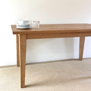 May include: A rectangular wooden dining table with four tapered legs and a natural light brown wood grain finish. On the surface of the table sits a stack of three white ceramic plates, a white bowl, and two clear glass tumblers with a textured pattern.