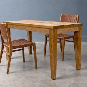 May include: A rectangular wooden dining table with four straight legs and two matching chairs. Each chair features a light brown wooden frame and a seat and backrest constructed from woven brown leather strips in a grid pattern.