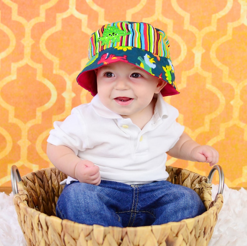 May include: A baby boy wearing a white shirt, blue jeans, and a colourful bucket hat with a dinosaur print. The baby is sitting in a woven wicker basket.