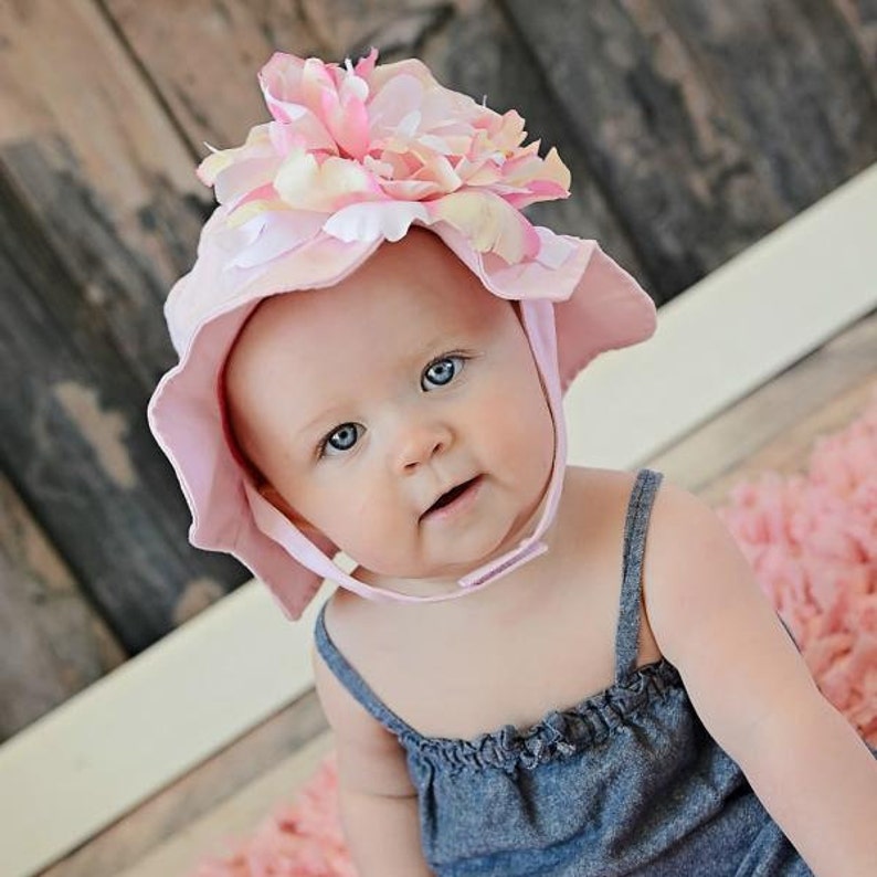 May include: A baby girl wearing a pink sun hat with a large pink flower on top. The hat has a wide brim and ties under the chin. The baby is wearing a denim dress.