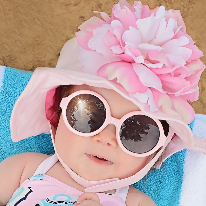 May include: A baby wearing a pink sun hat with a large pink flower on top and round pink sunglasses. The baby is lying on a blue and white striped towel.