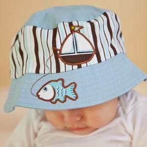 May include: A light blue baby sun hat with a brown and white striped band. The hat has an embroidered sailboat and a fish with a fishing line.