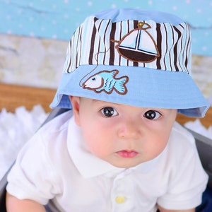 May include: A baby wearing a blue and white striped bucket hat with a sailboat and fish embroidery. The hat has a brown brim and a blue crown.