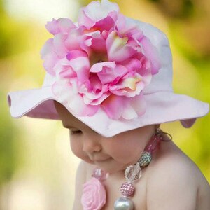 May include: A baby girl wearing a pink sun hat with a large pink flower on top. The hat has a wide brim and is made of a soft fabric. The baby is also wearing a pink and white beaded necklace.