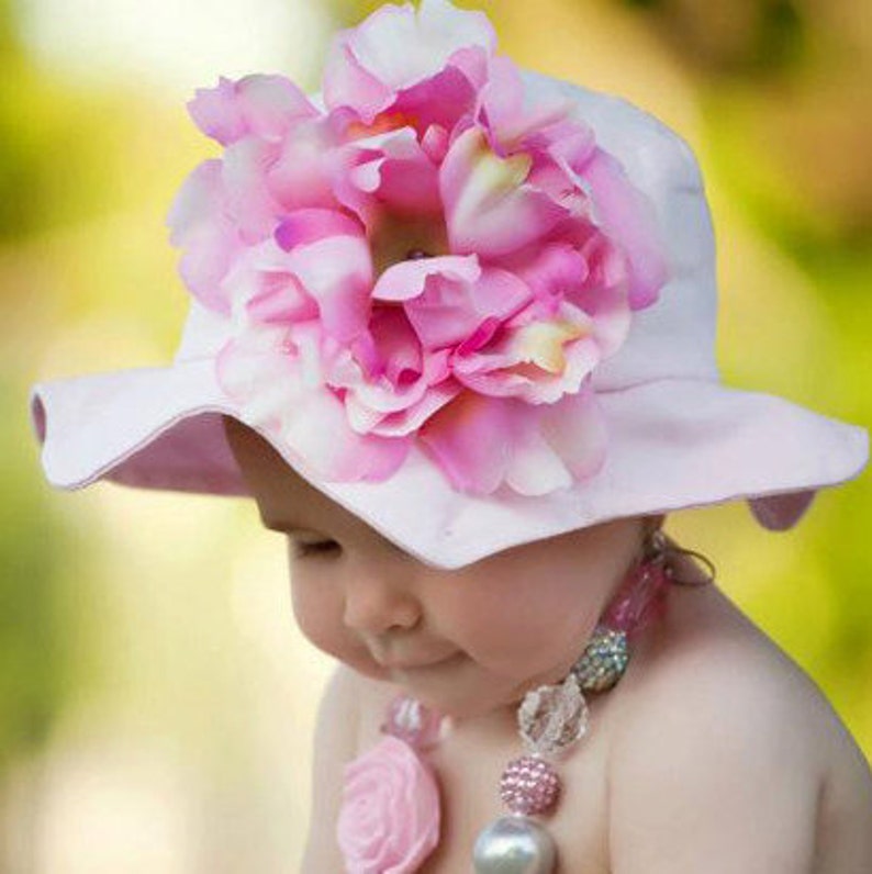 May include: A baby girl wearing a pink sun hat with a large pink flower on top. The hat has a wide brim and is made of a soft fabric. The baby is also wearing a pink and white beaded necklace.