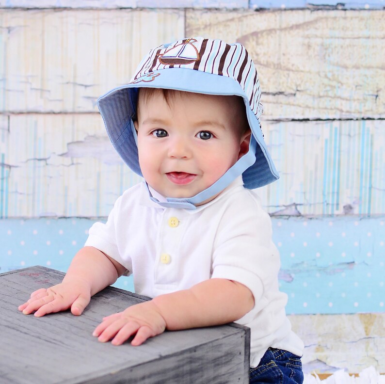 May include: A baby wearing a blue and white striped sun hat with a sailboat design. The hat has a chin strap and is made of a soft, breathable fabric. The baby is sitting on a wooden box and looking at the camera.