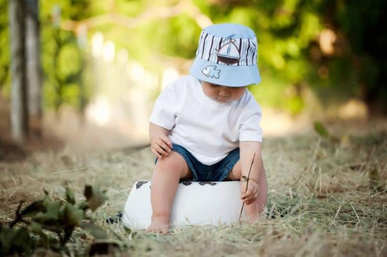 May include: A young child wearing a blue and white striped hat with a sailboat design sits on a white stool. The hat has a fish design on the front. The child is wearing a white t-shirt and blue denim shorts.