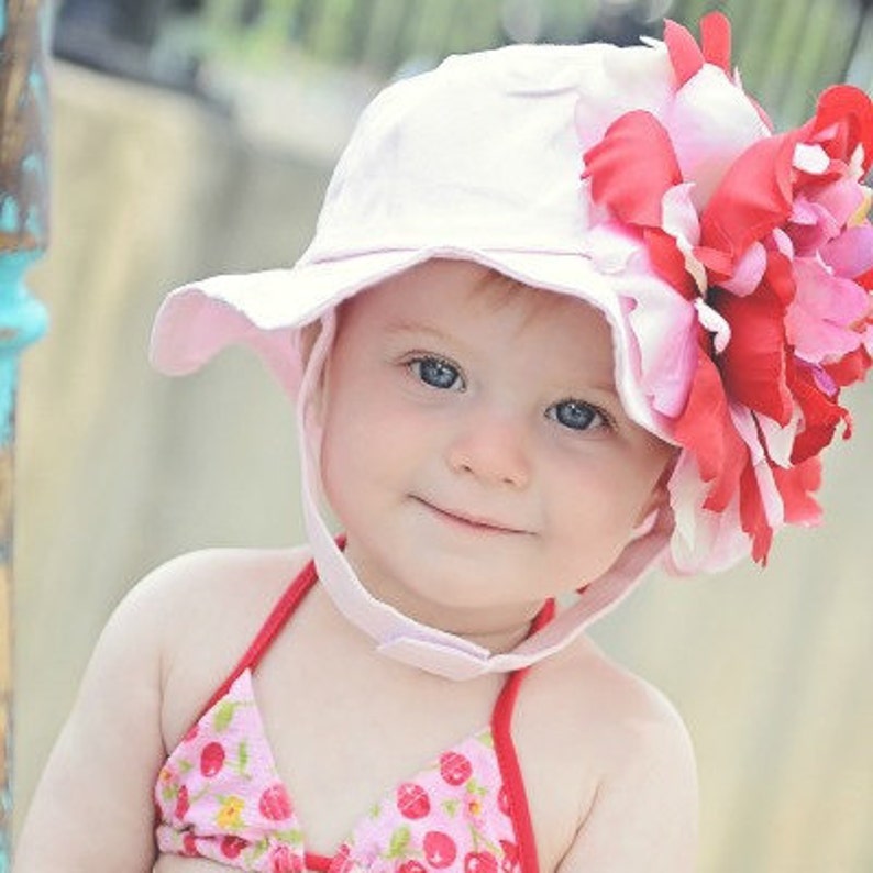 May include: A baby girl wearing a pink sun hat with a large red and pink flower on the side. The hat has a chin strap and the baby is smiling.