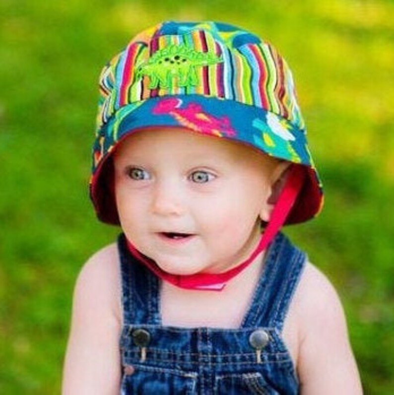 May include: A baby wearing a blue and green sun hat with a dinosaur design. The hat has a chin strap and is made of a soft, breathable fabric.