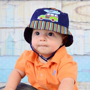 May include: A baby wearing a blue and white striped sun hat with a car and surfboard design. The hat has a chin strap. The baby is wearing an orange polo shirt.