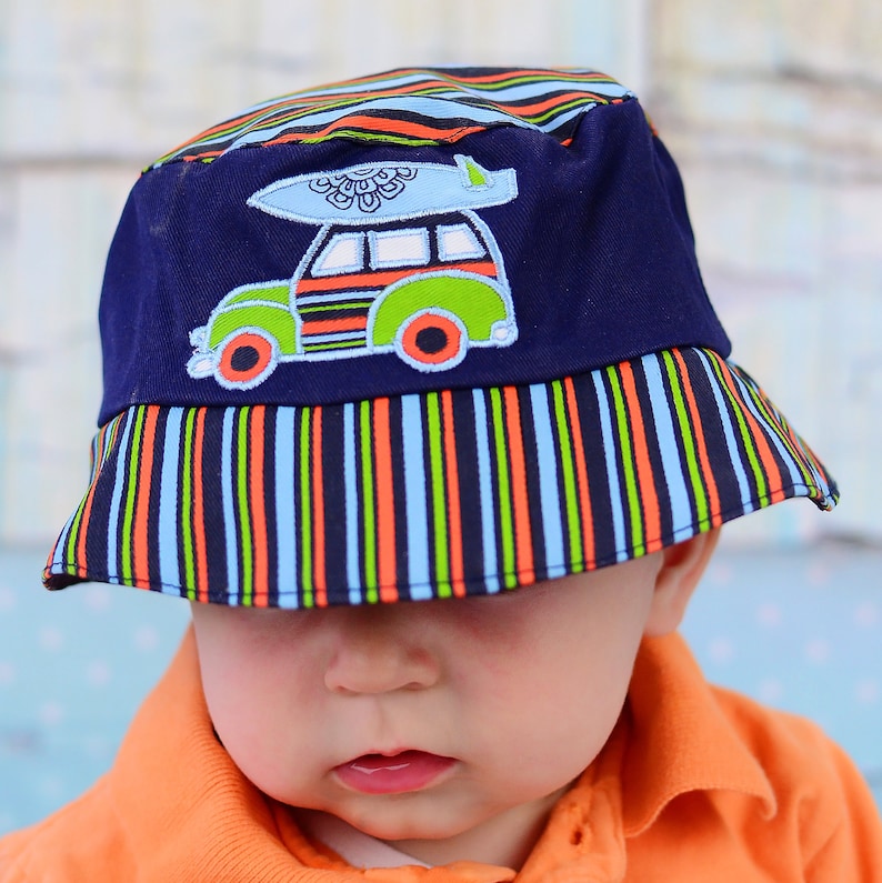 May include: A blue and white striped bucket hat with a colourful cartoon car and surfboard design. The hat is worn by a child wearing orange clothing.